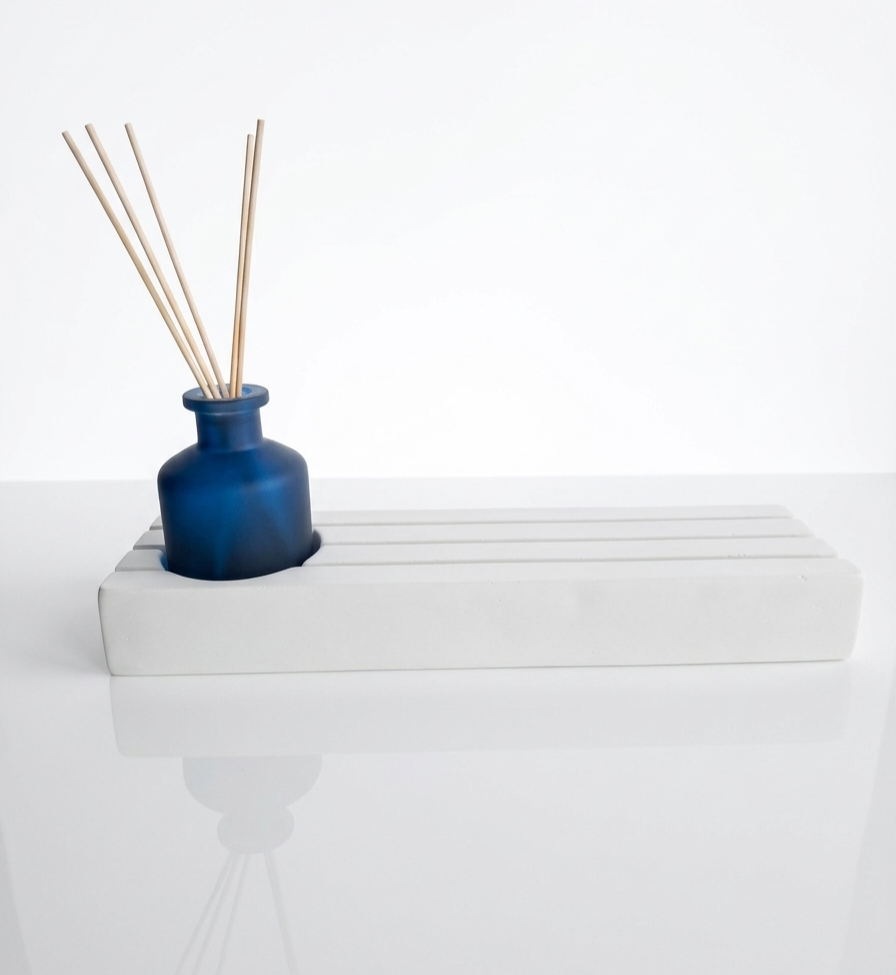 Minimalistische witte houder met een diepblauw glazen vaasje en geurstokjes op een glanzende witte ondergrond.