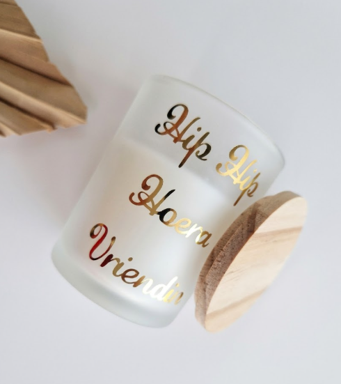 Luxe matglazen kaars met gouden tekst 'Hip Hip Hoera Vriendin' en houten deksel, gefotografeerd van bovenaf op een witte achtergrond.