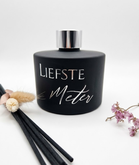 Mat zwart glazen flesje met zilveren geurstokjes en de tekst 'Liefste Meter', gedecoreerd met droogbloemen.