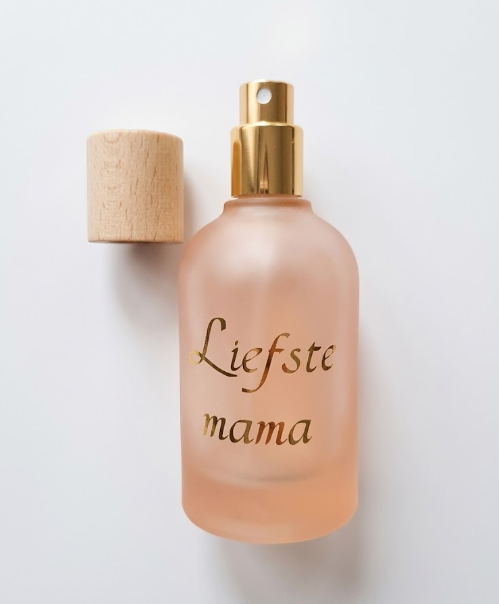Matroze glazen parfumfles met gouden opdruk 'Liefste mama' en een houten dop op een witte achtergrond.