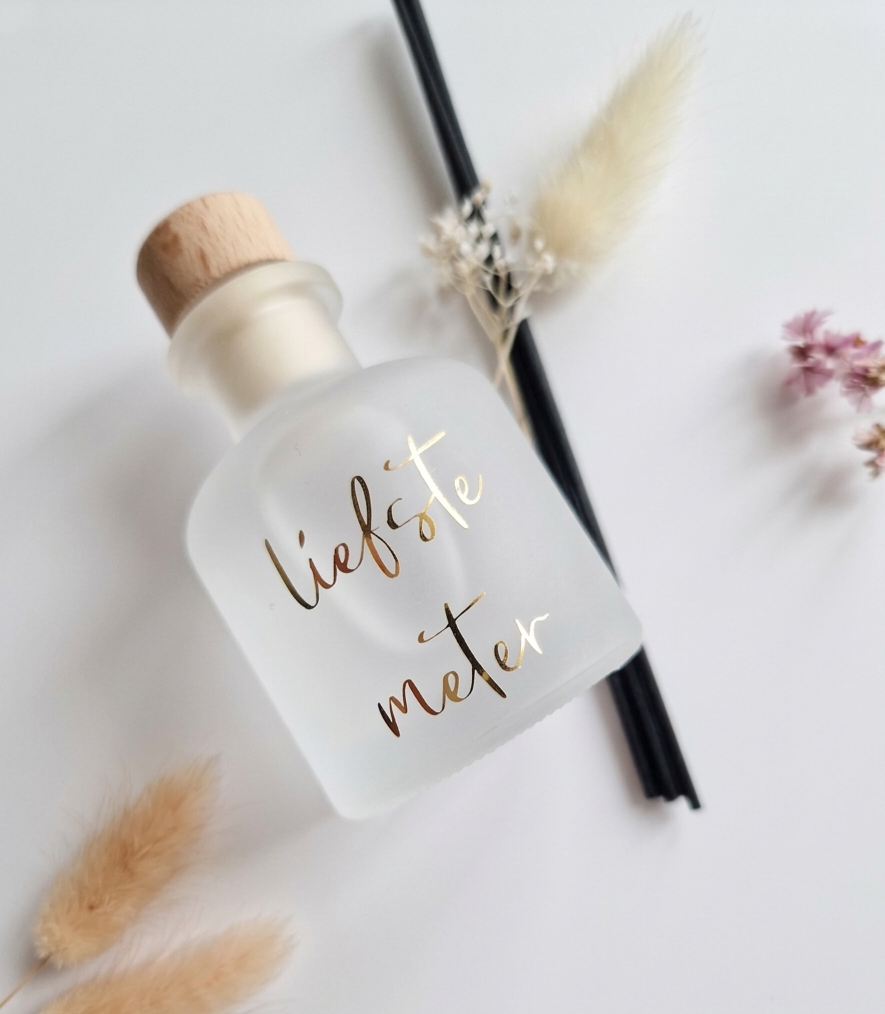 Mat wit glazen flesje met gouden belettering 'Liefste meter', een houten dop en zwarte geurstokjes met droogbloemen.