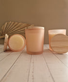 Twee sfeervolle geurkaarsen in perzik kleurig matglas met een natuurlijk houten deksel, gepresenteerd op een lichte houten ondergrond met een gedroogd palmblad op de achtergrond.