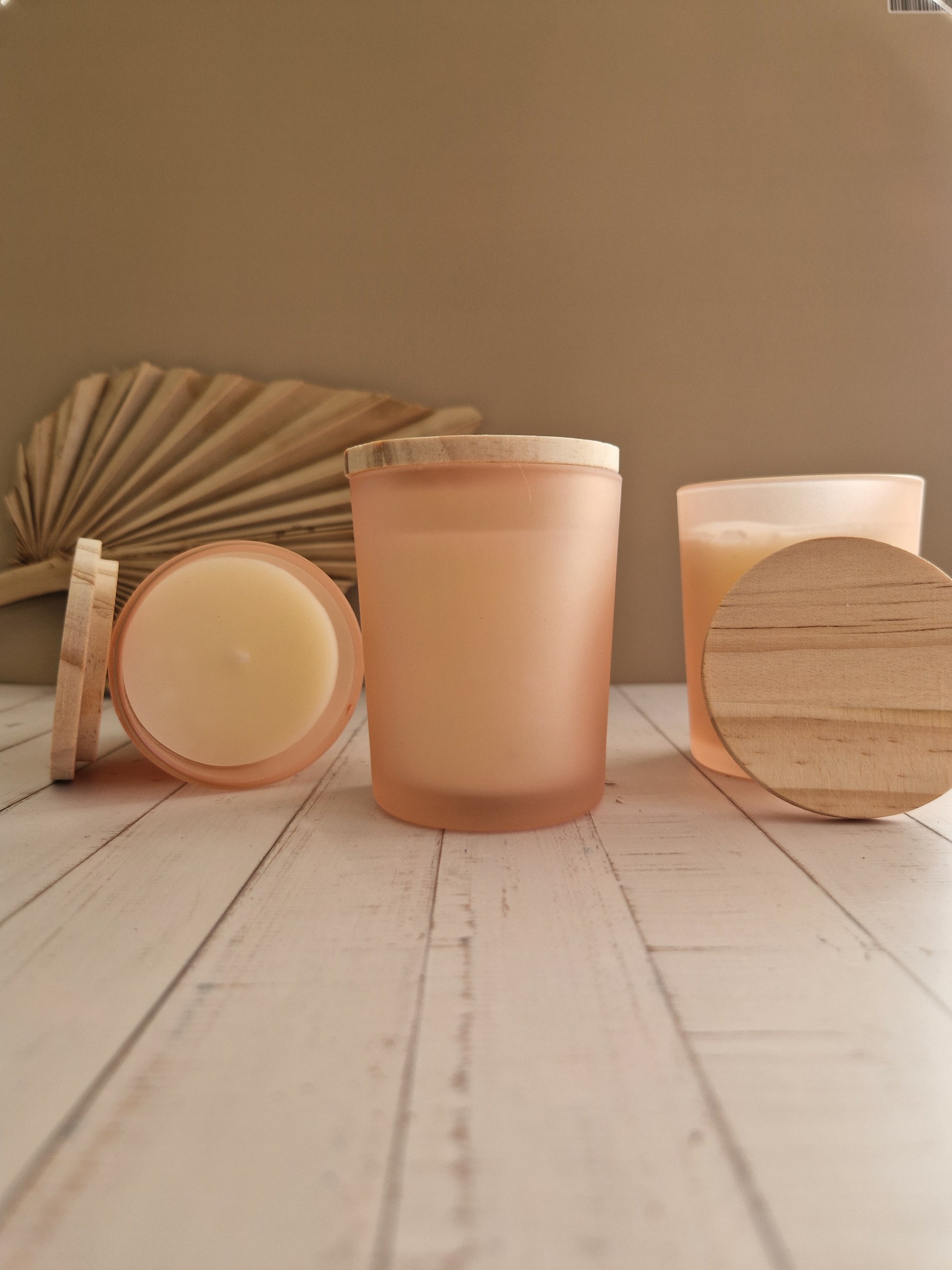 Twee sfeervolle geurkaarsen in perzik kleurig matglas met een natuurlijk houten deksel, gepresenteerd op een lichte houten ondergrond met een gedroogd palmblad op de achtergrond.