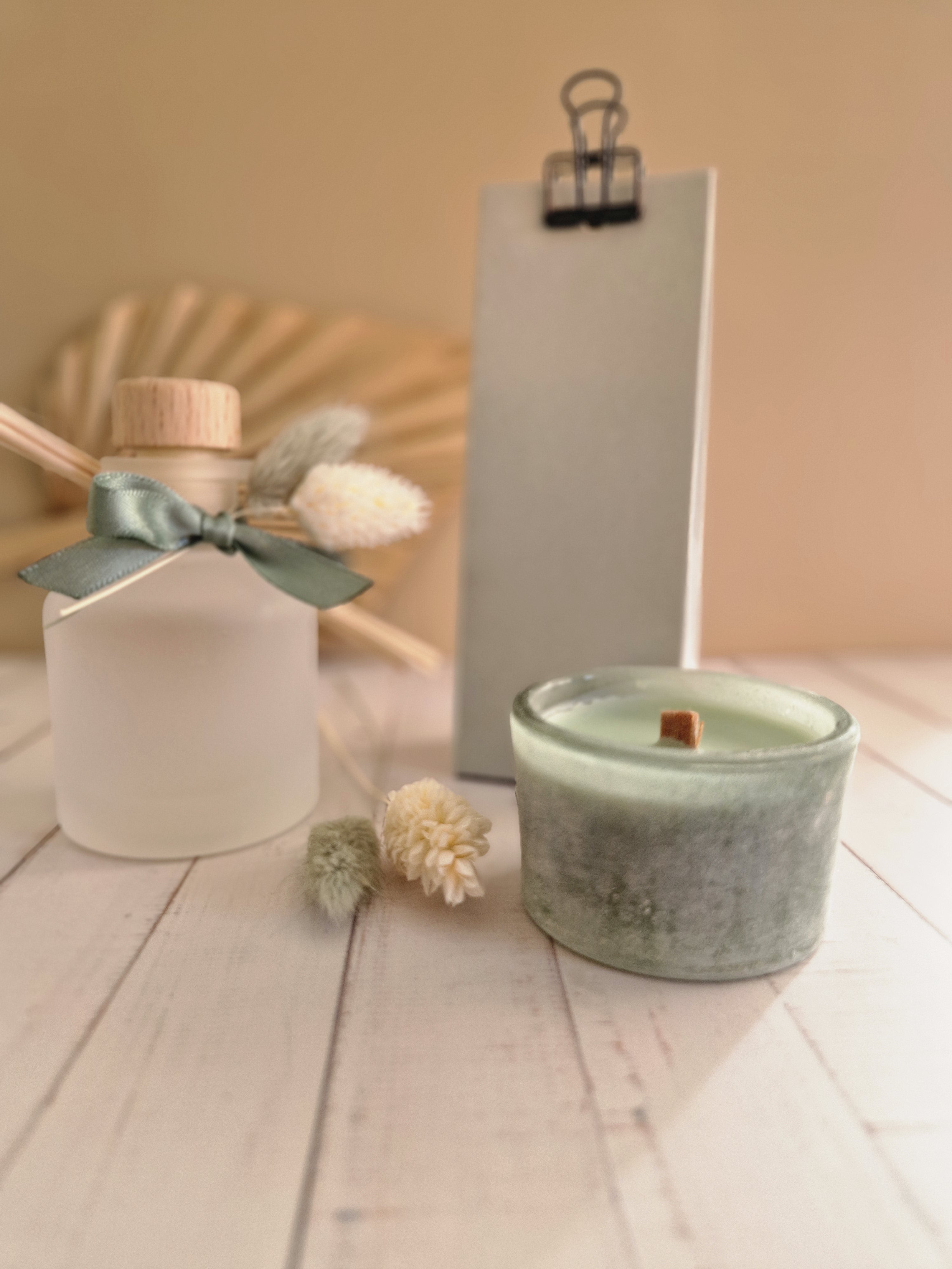 Een minimalistische lifestyle set bestaande uit een matglazen diffuser met houten dop en een saliegroene kaars met houten lont, gedecoreerd met droogbloemen en een groen strikje op een houten ondergrond.