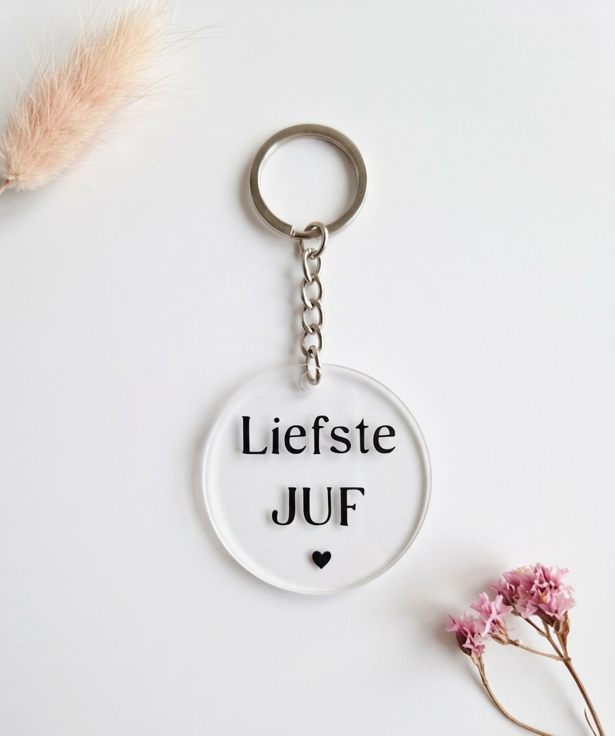 Ronde transparante sleutelhanger met de tekst Liefste Juf en een zwart hartje, geflankeerd door droogbloemen op een witte achtergrond.