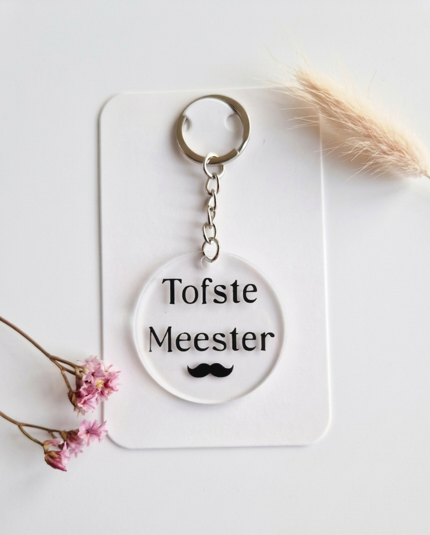 Ronde transparante acryl sleutelhanger met de tekst 'Tofste Meester' en een zwarte snor icoon op een witte achtergrond.