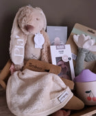 Een kartonnen cadeaumand gevuld met babyproducten, waaronder een beige knuffelkonijn met de naam 'Pia' op het oor, een Timboo slabber, BIBS fopspenen en een Liewood drinkbeker.