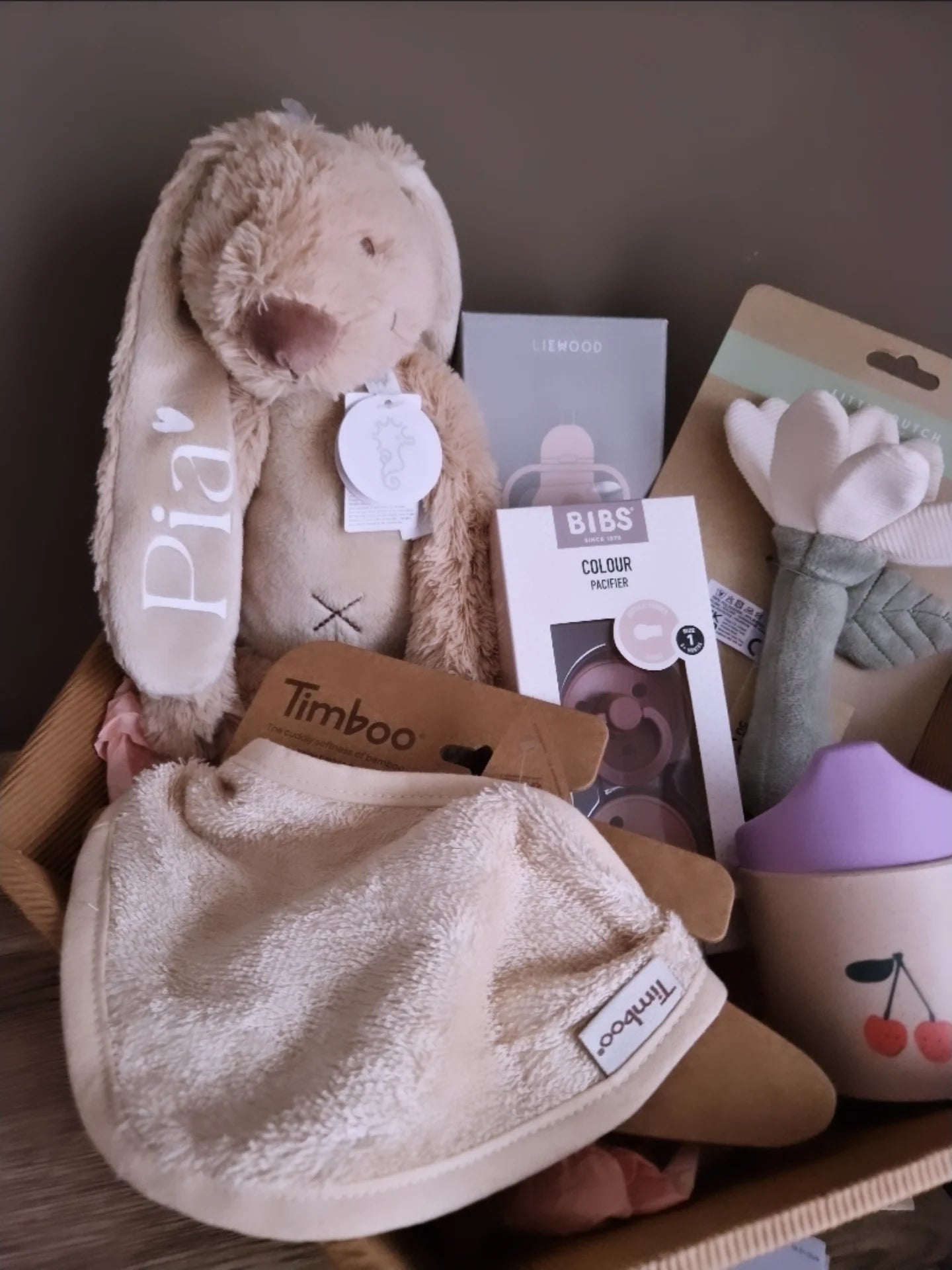Een kartonnen cadeaumand gevuld met babyproducten, waaronder een beige knuffelkonijn met de naam 'Pia' op het oor, een Timboo slabber, BIBS fopspenen en een Liewood drinkbeker.