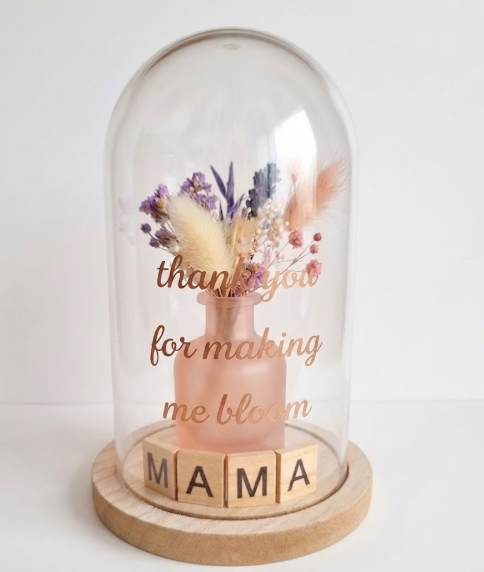 Glazen stolp met houten voet, gevuld met een roze vaasje met droogbloemen en houten letterblokjes die 'MAMA' spellen, met de gouden tekst 'thank you for making me bloom' op het glas tegen een witte achtergrond.